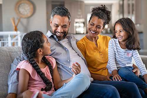 Image of a multi cultural family with a mom, dad, and two kids smiling at each other sitting on a couch